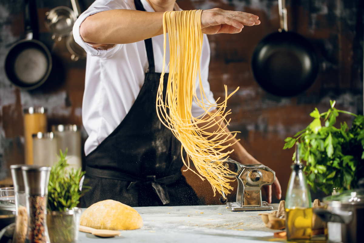 Chef making spaghetti noodles with pasta machine on kitchen table with some ingredients around.